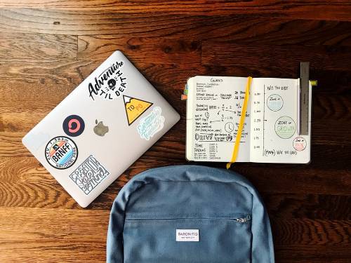 a back pack and laptop with decorative stickers and a book ontop of a table