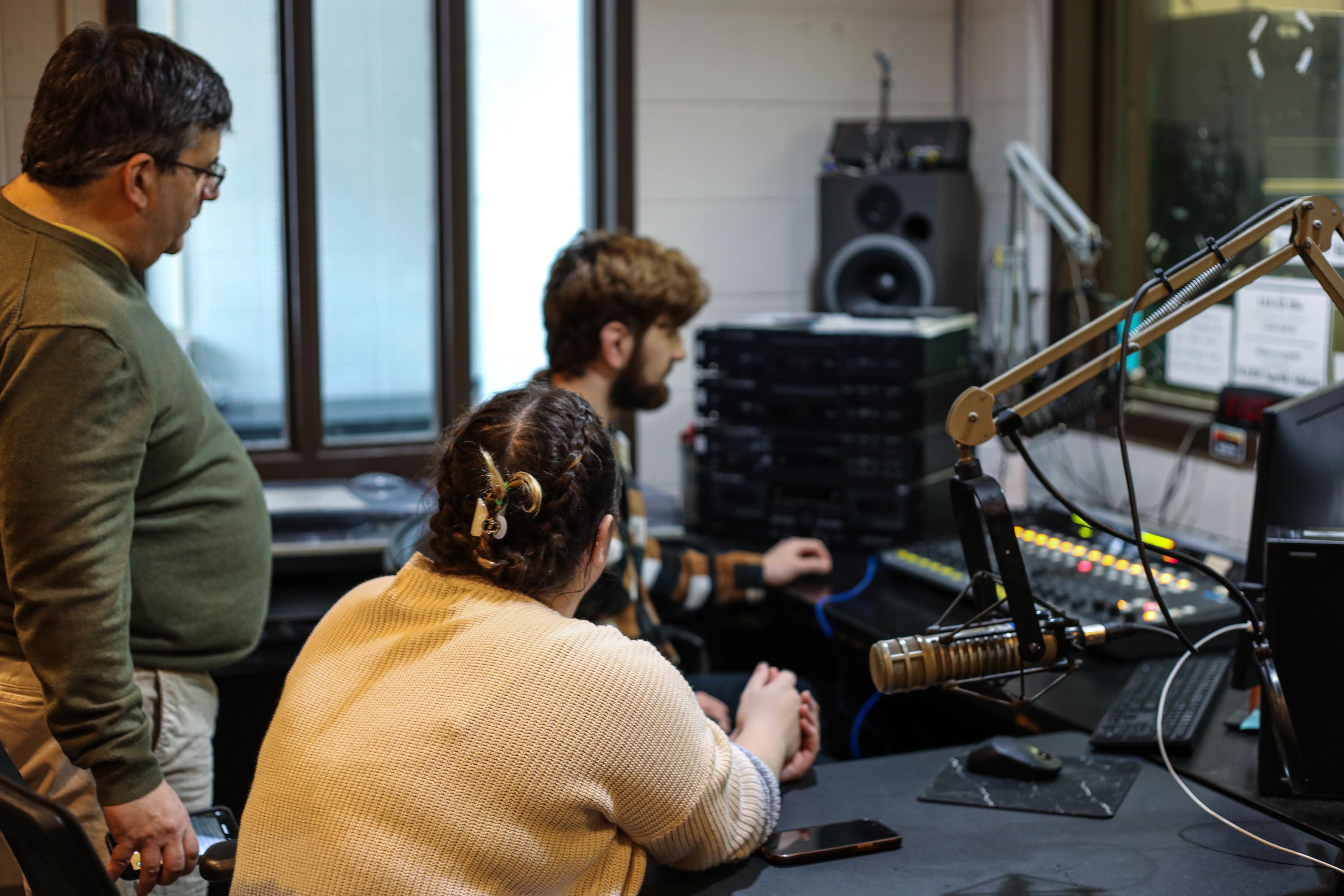 Three students sitting in a radio studio with mics and switch boards around them. Prepping to go live on air. 