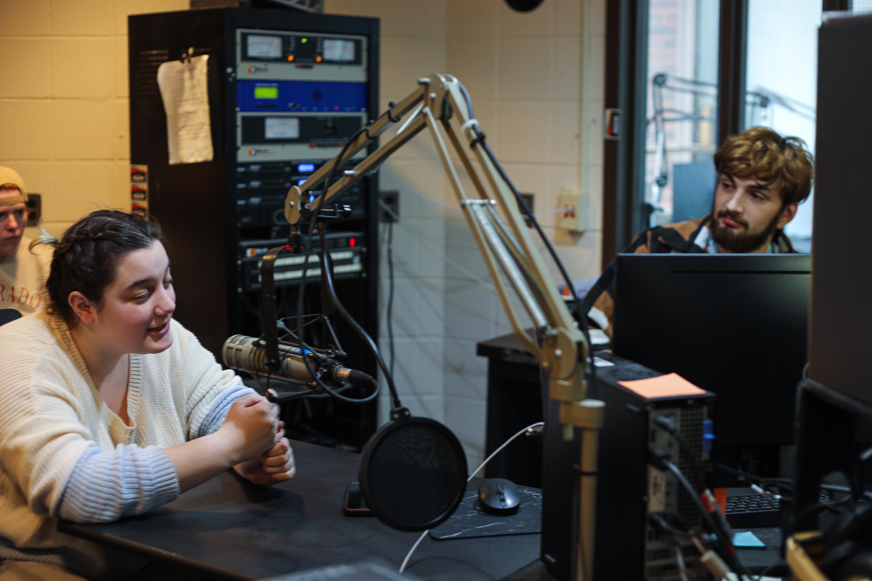 Students in radio station preparing to go on air. Female student sits in front of a mic. 