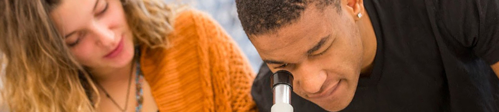 Student looking through a microscope while his lab partner takes notes